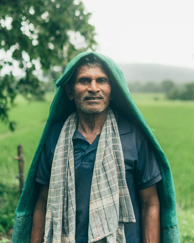 A farmer in rain-cloth, contemplative — knowledge in the field, weathered by it.